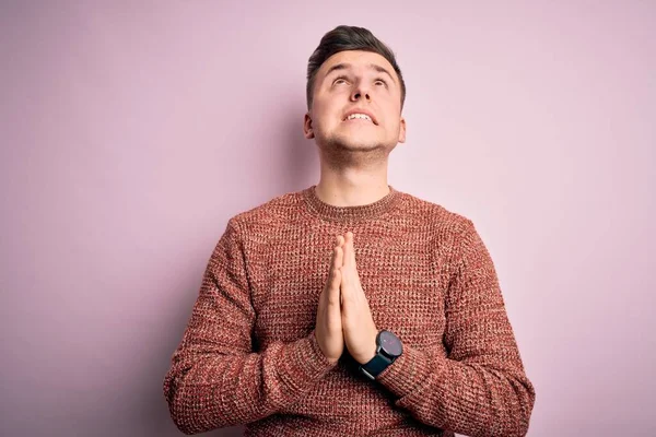 Young handsome caucasian man wearing casual winter sweater over pink isolated background begging and praying with hands together with hope expression on face very emotional and worried. Begging.