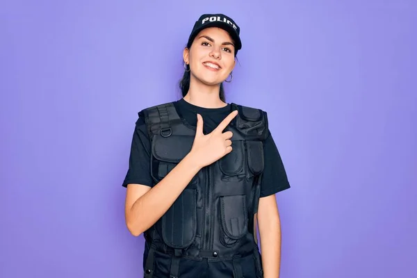 年轻的女警察身穿防弹背心制服 穿着紫色背景的制服 面带微笑 手指手画脚地朝侧面看去 脸上洋溢着愉快而自然的表情 — 图库照片