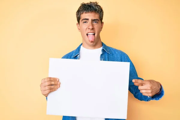 Young Handsome Man Holding Blank Empty Banner Sticking Tongue Out — Stock Photo, Image