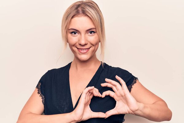 Молодая красивая блондинка в повседневной футболке стоит на изолированном белом фоне улыбаясь в любви показывая символ сердца и форму с руками. Романтическая концепция.