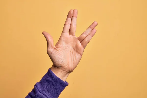 Hand of caucasian young man showing fingers over isolated yellow background greeting doing Vulcan salute, showing hand palm and fingers, freak culture