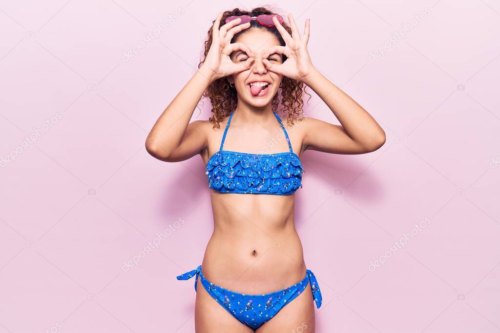 Beautiful kid girl with curly hair wearing bikini and sunglasses doing ok gesture like binoculars sticking tongue out, eyes looking through fingers. crazy expression. 