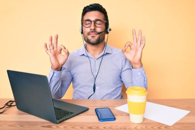 Çağrı merkezindeki operatör kulaklığı takmış, gözleri kapalı, meditasyon yaparken parmaklarıyla gülümseyen İspanyol genç bir adam. yoga konsepti. 