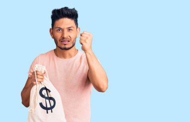 Elinde dolar sembolü olan yakışıklı Latin Amerikalı genç adam öfkeyle bağırıyor, öfkeyle bağırıyor ve el kaldırıyor. 