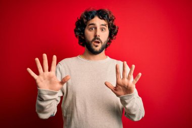 Sakallı genç yakışıklı bir adam kırmızı arka planda dikiliyor. Korkudan dehşete düşmüş ve korkmuş. El hareketlerini durdur, şok içinde bağır. Panik konsepti.