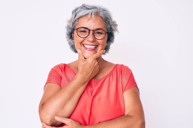 Son sınıf Latin kökenli, gri saçlı, günlük kıyafetler ve gözlük takan, kameraya kendinden emin bir şekilde bakan, kollarını kavuşturup çenesine kaldırmış gülümseyen kadın. Olumlu düşünmek. 