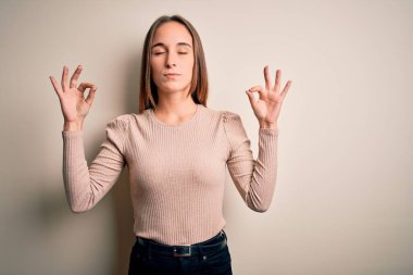 İzole edilmiş beyaz arka plan üzerinde duran sıradan bir süveter giyen genç ve güzel bir kadın rahatla ve parmaklarıyla meditasyon hareketi yaparken gözleri kapalı gülümse. Yoga konsepti.