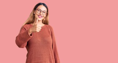 Gündelik kıyafetler ve gözlük takan genç ve güzel bir kadın elleriyle mutlu bir jest yapıyor. Kameraya bakıp başarıyı gösteren ifade. 