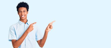 Genç Afro-Amerikalı sıradan kıyafetler giyen, gülümseyen ve kameraya bakan iki elini ve parmağını yana doğru gösteren bir adam.. 