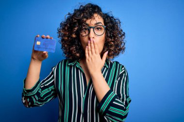 Genç, kıvırcık, gözlüklü bir kadın. Ödeme için kredi kartı parası tutuyor. Ağzını kapatıyor.