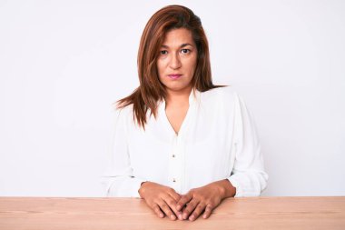 Orta yaşlı esmer, Latin iş kadını, gündelik beyaz gömlekli, masada oturan, şüpheci ve sinirli, sorun yüzünden üzgün. negatif kişi. 