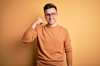 Genç, beyaz, gözlüklü ve sarı arka planda sıradan bir süveter giyen yakışıklı adam telefonla el ve parmaklarıyla telefonla konuşuyor gibi gülümsüyor. İletişim kavramları.