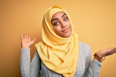 Genç ve güzel Afro-Amerikan kızı, izole edilmiş sarı arka plan üzerine Müslüman başörtüsü takıyor. Kolları ve elleri havada, kafası karışmış bir ifade takınıyor. Şüphe kavramı.