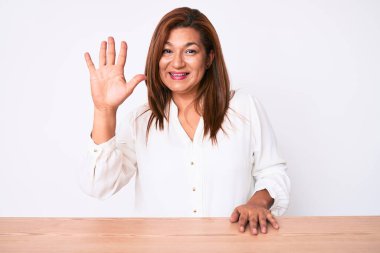 Orta yaşlı esmer, İspanyol iş kadını. Masada oturan ve parmaklarını 5 numarayı göstererek gülümseyen, kendinden emin ve mutlu bir kadın.. 