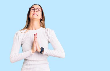 Beautiful brunette young woman wearing casual white sweater and glasses begging and praying with hands together with hope expression on face very emotional and worried. begging.  clipart