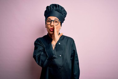 Orta yaşlı esmer aşçı kadın aşçı üniforması giyiyor ve şapka takıyor pembe arka planda el ağızda gizli dedikodular anlatıyor, kötü niyetli konuşmalar fısıldıyor.