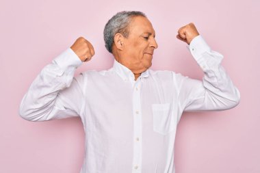 Soyutlanmış pembe arka planda zarif gömlekler giyen yaşlı, gri saçlı adam gururla gülümseyen kol kaslarını gösteriyor. Fitness konsepti.