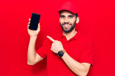 Sakallı, teslimat üniforması giyen, elinde akıllı telefon tutan, mutlu, eli ve parmağı olan genç yakışıklı bir adam. 