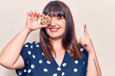 Elinde çikolatalı kurabiye tutan ve gülümseyen genç ve cüsseli bir kadın bir fikir veya soruyla mutlu bir yüzle işaret ediyor. 