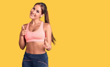 Spor kıyafetleri giymiş genç ve güzel bir İspanyol kadın mutlu ve komik yüzüyle kamerayı işaret ediyor. iyi enerji ve titreşimler. 