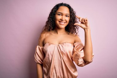 Kıvırcık saçlı, pembe arka planda sıradan tişörtler giyen, el hareketleriyle küçük beden işareti yapan, gülümseyen ve kameraya bakan genç ve güzel bir kadın. Ölçü kavramı.