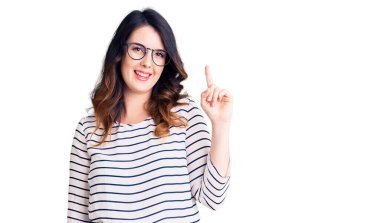 Güzel, genç, esmer, günlük kıyafetler ve gözlük takan bir numaralı parmağını gösterip işaret eden, kendinden emin ve mutlu bir şekilde gülümseyen kadın.. 