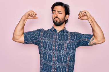 Pembe arka planda hippi tarzı giyinen yakışıklı İspanyol bohem adam gururla gülümseyen kol kaslarını gösteriyor. Fitness konsepti.