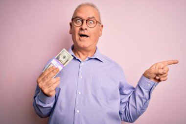Orta yaşlı, ağarmış bir adam, soyutlanmış pembe arka planda bir deste banknot tutuyor. Elleri ve parmakları kenarda mutlu mesut işaret ediyor.