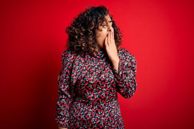 Genç ve güzel kıvırcık Arap kadın kırmızı arka planda dikilen gündelik çiçekli elbiseler giyiyor. Ağzı eliyle kapatmaktan sıkılmış. Huzursuz ve uykulu..