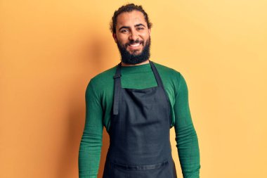 Genç Arap adam berber önlüğü takıyor. Pozitif ve mutlu görünüyor. Duruyor ve gülümsüyor. 