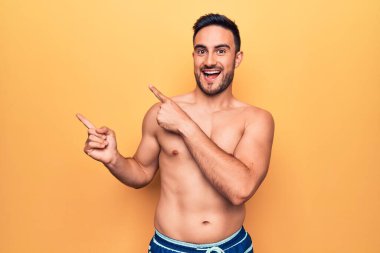 Sakallı, kolsuz tişörtlü, sarı arka planda duran, gülümseyen ve kameraya bakan genç, yakışıklı bir adam..