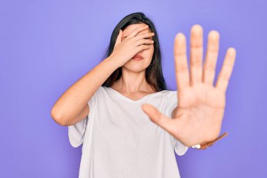 Genç, güzel, esmer bir kadın mor arka plan üzerine günlük beyaz tişört giyip gözleri ellerle kaplıyor ve üzgün ve korku ifadeleriyle jest yapıyor. Utanç verici ve olumsuz bir kavram..