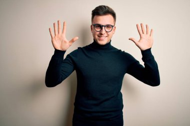 Gözlüklü, sıradan bir süveter giyen genç, yakışıklı beyaz bir adam izole edilmiş arka planda on numarayı işaret ederken kendinden emin ve mutlu bir şekilde gülümsüyor..
