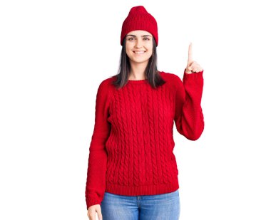 Süveter ve yün şapka giyen genç ve güzel bir kız bir numaralı parmağını gösterip işaret ederken kendinden emin ve mutlu bir şekilde gülümsüyor.. 