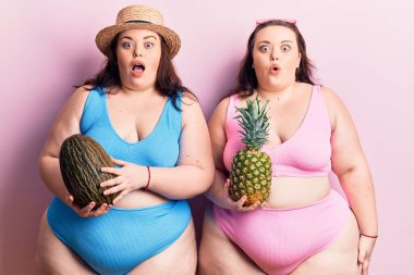 Bikini giyen, elinde kavun ve ananas tutan genç ve büyük beden ikizler. Şaşırmak için ağzı açık kalmış, inançsız bir yüz. 