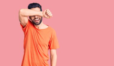 Sakallı, sıradan tişörtler giyen, gözleri kollarıyla kaplayan neşeli ve komik gülümseyen genç yakışıklı bir adam. kör kavram. 