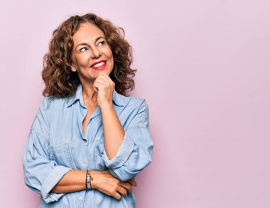 Orta yaşlı güzel bir kadın pembe arka planda dikilmiş sıradan bir kot gömlek giyiyor ve elleri çenesinde soru soruyor, düşünceli bir yüz ifadesi var. Gülen ve düşünceli bir yüz. Şüphe kavramı.