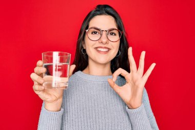 Genç ve güzel bir kadın izole edilmiş kırmızı arka planda bir bardak temiz su içiyor. Tamam işareti yapıyor, mükemmel bir sembol.