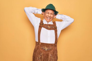 Kıdemli gri saçlı adam sarı arka planda geleneksel Alman Ekim Festivali kıyafeti giyiyor. Kollarını ve ellerini başının ve boynunun arkasına koyup gülümsüyor.