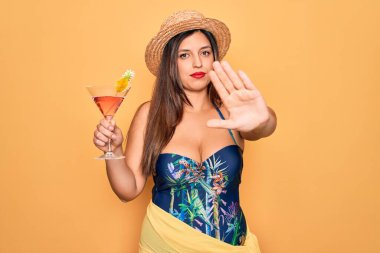 Yaz mayosu giyen genç İspanyol kadın sarı arka planda kokteyl içiyor. Açık elle dur işareti yapıyor. Ciddi ve kendinden emin bir ifadeyle, savunma hareketi yapıyor.