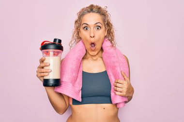 Güzel, sarışın, spor yapan, havlu tutan, protein içeceği ile titreyen, ağzı açık, ağzı açık, şaşırmış, inançsız bir kadın.