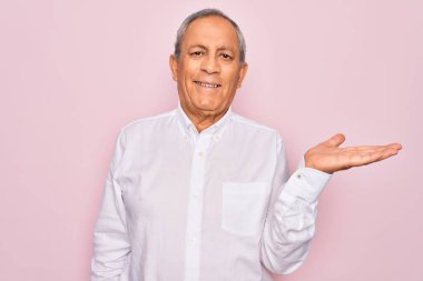 Üst düzey yakışıklı, gri saçlı, zarif gömlekli, pembe arka planda, neşeli bir şekilde gülümseyen ve avuç içi kameraya bakan bir adam..