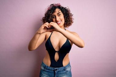 Genç, güzel bir Arap kadın, mayo ve pembe arka planda güneş gözlüğü takarak tatilde. Aşkla gülümsüyor. Elleriyle kalp sembolü ve şekli gösteriyor. Romantik konsept.