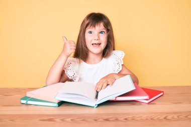 Küçük beyaz kız, uzun saçlı okul sınavına çalışıyor mutlu ve pozitif gülümsüyor, başparmağıyla mükemmel ve onay işareti yapıyor. 