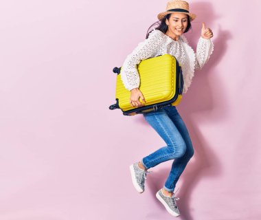 Tatilde olan genç ve güzel bir kadın. Yazlık kıyafetler ve mutlu bir şekilde gülümseyen bir şapka. Yüzünde tebessümle kabin çantasını izole edilmiş arka planda tutarken