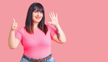 Genç ve cüsseli kadın gündelik giysiler giyiyor. Altıncı parmaklarını gösterip işaret ederken kendinden emin ve mutlu gülümsüyor.. 