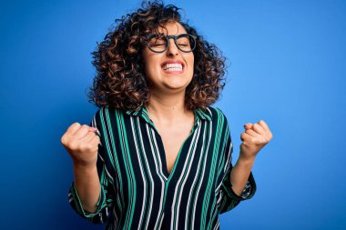 Mavi arka planda çizgili gömlek ve gözlük takan güzel, kıvırcık Arap kadın. Kolları havada, gözleri kapalı zaferi kutlarken gülümsüyor. Kazanan konsept.