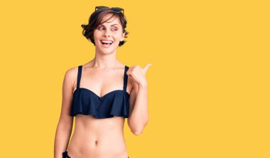 Kısa saçlı, bikinili, mutlu yüzüyle gülümseyen ve başparmağıyla yan tarafı gösteren güzel bir kadın.. 