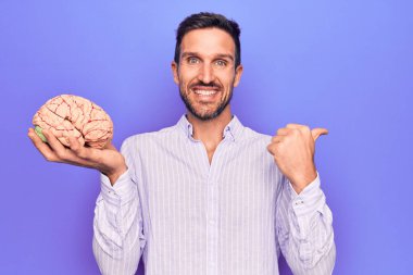 Genç yakışıklı adam hafızasını istiyor beyni izole edilmiş mor arka planda tutuyor başparmağıyla yan tarafa işaret ediyor mutlu bir şekilde ağzı açık gülümsüyor.