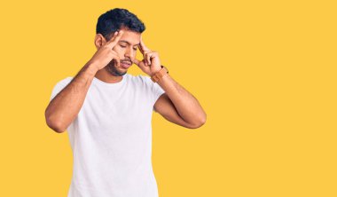 Elleri başının üstünde, gündelik kıyafetler giyen genç latin adam, stres yüzünden baş ağrısı. migren ağrısı. 